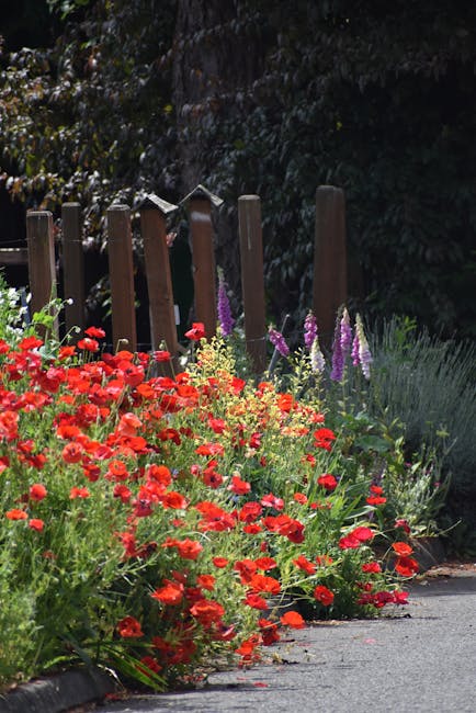 A lush garden scene featuring a variety of plants, shrubs, and trees. In the foreground, there are vibrant pink and red flowering bushes alongside ornamental grasses with cream and green striped leave